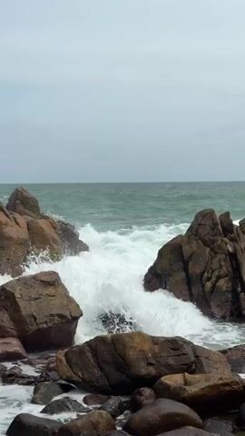 The video shows a rocky coastline with waves crashing against large boulders. The water is a greenish-blue, and the sky is overcast with gray clouds. The waves are powerful, creating white foam as they hit the rocks. The foreground is filled with smaller stones and pebbles. The overall impression is one of a wild and untamed natural landscape.