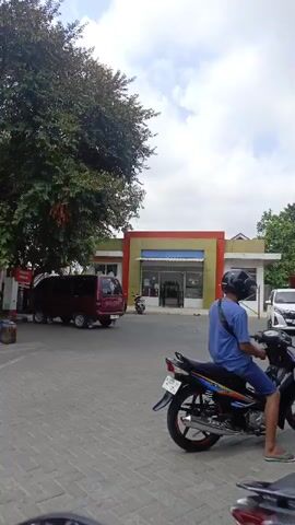 A medium shot shows a person sitting on a black motorcycle. The person is wearing a blue t-shirt and blue shorts, and a black helmet. The motorcycle has a white license plate and red accents on the wheels. The background shows a building with a yellow and orange facade, and trees. There are other vehicles visible in the background, including a red van and a white car. The ground is paved with gray bricks. The sky is cloudy.