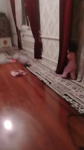 The video shows a section of a room with a long, patterned carpet and heavy red curtains. White lace curtains are also visible next to the red ones. The camera focuses on the curtains and the floor. A wooden table is visible in the foreground on the left side. A child’s scream can be heard.