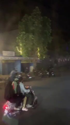At night, a light-colored scooter carrying two people travels on a dark road. The scooter has a red taillight. The person driving is wearing a light-colored shirt. The passenger in the back has a large black backpack. The road is lined with trees and buildings. Another scooter goes past in the opposite direction.