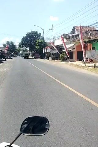 The video shows a street scene from the perspective of someone riding a motorcycle. The road is paved with a yellow line down the center. On the right side of the road, there are buildings with red and white flags hanging outside. A yellow truck is driving away from the camera on the left side of the road. Further down the road, there are more buildings and trees. The sky is blue and clear. A black car is parked on the right side of the road. A red scooter is also visible on the right side of the road. Shadows of trees are cast on the road.