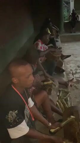 A group of men are gathered in what appears to be an outdoor gathering space. Six individuals are visible, seated along a low wall. One man, closest to the camera, is holding a small bundle of what looks like palm leaves and using them to strike a hollow log, creating a percussive sound. Another man further down the line is playing a hand drum, adding to the rhythmic music. The remaining men are either watching or participating in the music-making in some other way. In the background, another man can be seen sitting on a higher wall. The environment seems informal and relaxed. The lighting is dim, suggesting it may be evening.