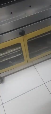 The video shows an inside view of an oven. Several trays of pastries are visible. Some appear to be round and sprinkled with sesame seeds, while others are triangular and layered. The oven has a yellow exterior with glass doors. The interior is metal with visible heating elements. The overall scene suggests a commercial bakery or kitchen environment.