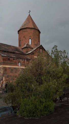 The video shows an exterior view of the Saghmosavank Monastery in Armenia on a cloudy day. The camera pans across the complex, showcasing its architecture, which includes a church with a red brick tower topped with a cross, a bell tower, and stone walls. The ground is paved with stone. Several people are walking around the courtyard, some with umbrellas. The environment is overcast, and the colors are muted. Vegetation, including trees and bushes, is visible around the buildings.