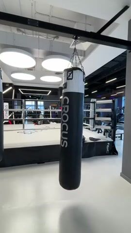 The video opens with a view of a punching bag hanging from a metal support in a gym. The bag is black with white lettering that reads "CROCUS". In the background, a boxing ring is visible. The camera then pans to the right, revealing a weight training machine and then moves further to show rows of treadmills and other exercise equipment. A person is seen walking on one of the treadmills in the distance. The environment appears to be a modern, well-lit fitness facility with a clean white floor and dark ceiling.