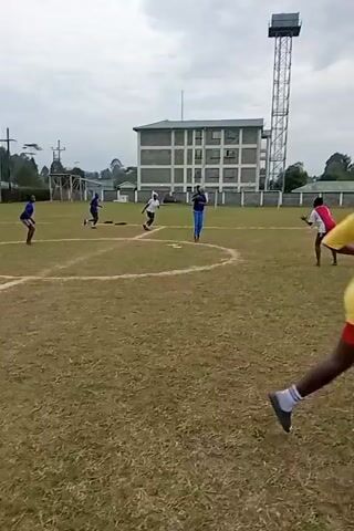 The video shows a group of people playing a game on a grass field. There are four people initially visible, one wearing blue pants and a blue shirt, two others wearing blue and white shirts, and one wearing a white and red shirt with black shorts. As the video progresses, more people come into view, including one wearing an orange shirt and another wearing a white shirt. The people are running around and throwing a ball to each other. The environment includes a building, a tall tower, trees, and a wall. The sky is overcast.
