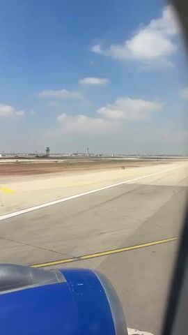 The video shows a view from an airplane window as it taxis on a runway. The foreground shows the blue and silver engine of the airplane. The middle ground shows a tarmac with white and yellow lines, indicating a runway. In the distance, there are airport buildings and a control tower under a blue sky with scattered white clouds. The plane appears to be moving slowly on the ground.