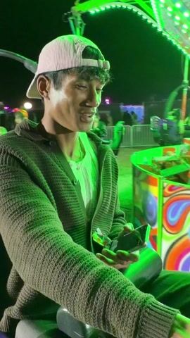 A young man with a baseball cap is riding an amusement park ride at night. He wears a grey cardigan and a white t-shirt. The ride has bright flashing lights and colorful psychedelic patterns. The man smiles at the camera briefly as the ride rotates. The background features other amusement park structures. The lighting on the scene shifts between green and blue.
