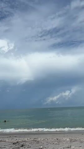 A low-angle, full-shot video of a beach with a person wading in the ocean. The sky is filled with dramatic clouds, with patches of blue sky visible and darker storm clouds gathering on the horizon. The ocean water is calm and a light green color, with small waves lapping at the shore. The sand in the foreground is light gray and appears wet near the water's edge. The person in the water is only visible from the shoulders up.