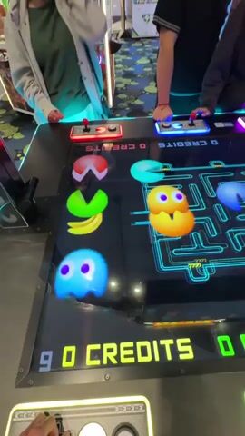 The video captures an engaging scene inside an arcade, specifically in Schaumburg, United States of America, where at least three individuals are actively playing a large, multi-player arcade game. The game is a modern, oversized version of Pac-Man, featuring a horizontally oriented screen that displays a glowing neon blue maze.

Multiple players are gathered around the game table, each interacting with their designated control panel. On the left, a player in a light-colored top uses a red-lit joystick and buttons. In the center, another person wearing a dark hoodie is focused on a pink/purple-lit control panel. To the right, a third player in a dark top manipulates a blue-lit joystick and buttons, while the arm of a potential fourth player is visible near a green-lit control panel on the far right.

The game screen is brightly illuminated, showcasing the iconic Pac-Man maze filled with white pellets, larger power pellets, and various colored ghosts (blue, red, orange, green, yellow, pink) alongside the classic yellow Pac-Man characters. The players are seen actively guiding their characters through the maze.

The control panels prominently display illuminated text such as "CREDITS" and "INSERT COIN" in green and yellow hues. Numerical scores or credit counts like "100" and "200" are also visible, along with "0 CREDITS" on one panel. The background reveals other blurred arcade machines, confirming the setting. The general atmosphere appears lively and exciting, with sounds of enthusiasm from the players.