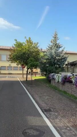The video shows a view from a street, looking towards a building with balconies adorned with flowers. The building is painted yellow and brown, with decorative paintings on its facade. In front of the building, there is a sidewalk paved with bricks and a planter filled with flowers. The street is asphalt, with a white line marking the edge. A few trees are visible on the left side of the street, as well as a smaller building in the background. The sky is blue with a few wispy clouds. The camera pans to the right, revealing more of the building and its surroundings. The overall impression is of a pleasant, well-maintained environment, likely a hotel or guesthouse in a scenic location.