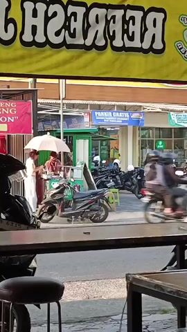 The video shows a street scene with several motorcycles and a car. In the foreground, there is a partial view of a table and a stool. In the background, there is a large yellow banner with white text that appears to read "REFRESH" upside down. Below the banner, there are several storefronts. One storefront has a pink banner with text. Another storefront has a blue sign that reads "PARKIR GRATIS," meaning free parking. Several people are visible, including individuals on motorcycles and a few walking. One person is holding a white umbrella. A white car is driving past. There are also several parked motorcycles. The overall environment appears to be a busy street in a commercial area.