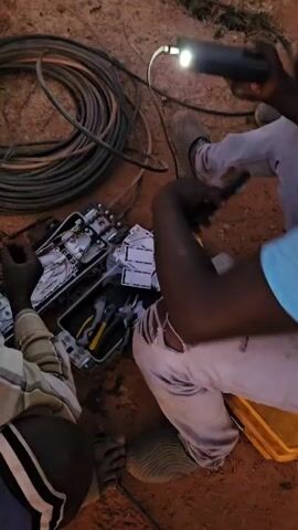 Two individuals are working on a fiber optic splicing job on a patch of red earth. A large coil of thick black cable lies to the upper left of the frame. One person is wearing a striped, long-sleeved shirt and blue shorts. The other wears torn white jeans and a blue t-shirt. They are kneeling on the ground, attending to two open splice boxes. Tools, including pliers, are visible within the boxes. The person in white jeans is using a black smartphone to photograph the work. A small yellow container sits near them. A light source, possibly a flashlight, illuminates the scene from above.