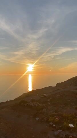 The video shows a golden sunset over the sea. The sun, a bright disc, is partially obscured by thin clouds. Its reflection creates a shimmering path of light on the water's surface. The sky is a blend of orange, yellow, and light gray. In the foreground, a rocky hillside is partially visible, with some debris scattered across it. There is also a small white vehicle parked on the hillside.