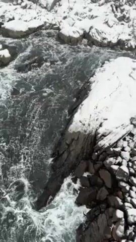 The video showcases a landscape of a seaside village amidst snowy conditions. Initially, the scene is focused on the rough sea water flowing around and between snow-covered rocks. As the view pans upwards, a series of red wooden houses appear along the coastline, all blanketed in snow. In the background, a large, snow-covered mountain looms, its peak shrouded in mist or fog. The overall weather appears to be snowy, with reduced visibility. The scene is tranquil, despite the rough sea.