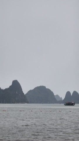 The video shows a body of water, likely a bay or sea, with several rocky, green islands in the distance. The sky is overcast and gray. In the foreground, a line of orange buoys floats on the water. A small boat with several people on board moves along the water in the middle ground.