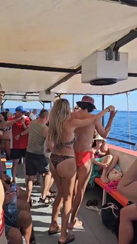 A group of people are on a boat party. Several individuals are dancing on the deck. In the foreground, a woman in a leopard-print bikini is dancing with a man wearing red swim briefs and a hat. Other people are visible in the background, some sitting on benches and others standing. The boat appears to be on the ocean, with water visible to the right. The scene is illuminated by natural light.