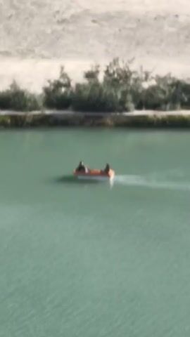 The video begins with a shot of a small orange boat with two people in it on a calm body of water. The boat is moving and creating a wake. In the background, there is a sandy bank with some trees. The camera then zooms out and pans up to reveal a wider view of the landscape. We see a large body of turquoise water with some patches of greenery growing in it. Beyond the water, there is a sandy area with several tents and vehicles, suggesting a campsite or a small settlement. Further in the background, a massive mountain range dominates the scene, with a waterfall cascading down its side. The sky is partly cloudy. The overall impression is of a serene and picturesque natural environment.