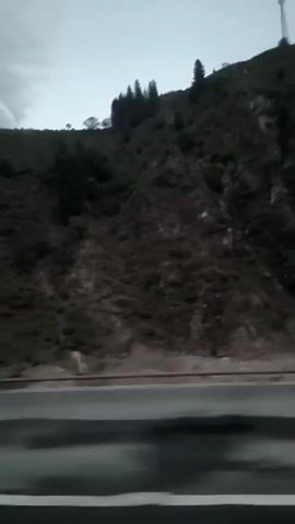 The video shows a view from a moving vehicle, possibly a car or bus. The camera is facing outwards, capturing the passing scenery. On the left side of the frame, a steep, tree-covered hillside is visible, with a road running alongside it. A light blue guardrail separates the road from the hillside. Further in the distance, the terrain appears to be mountainous and rocky. The sky is overcast, suggesting it may be dusk or a cloudy day. The image is slightly blurred due to motion, giving a sense of speed. Towards the right side of the frame, large cylindrical objects, possibly industrial spools or tanks, are visible on a flatbed trailer, also in motion. These objects are predominantly blue and white. The overall impression is that of traveling through a rural or mountainous area, possibly a highway.