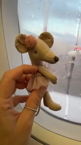 A person's hand, wearing a silver ring on their index finger, holds a small stuffed mouse by the window of an airplane. The mouse is beige with large ears and a pink pompom on its head, wearing a light pink tutu. Outside the window, an EasyJet airplane is parked at an airport gate. Several people in yellow safety vests are visible on the tarmac, along with a jet bridge and airport equipment. The window is streaked with water droplets. The sky appears cloudy and overcast.