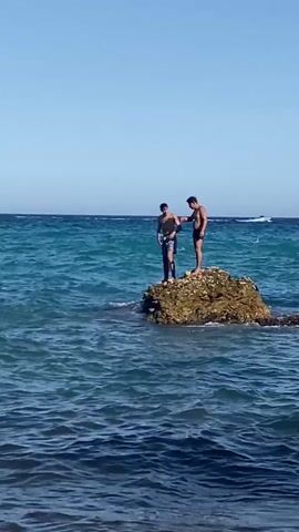 Two young males, appearing to be teenagers or in their early twenties, are standing on a large rock formation in the ocean. The male on the left is wearing patterned swim trunks, and the male on the right is wearing black swim trunks. Both are shirtless. The water is a vibrant blue with moderate waves. The sky is clear and blue. In the distance, a white boat or object is visible on the horizon. The rock appears to be covered in barnacles or small shells. The scene is captured from a low angle, looking up towards the rock and the individuals. The video footage is grainy, suggesting it was taken with a handheld device.