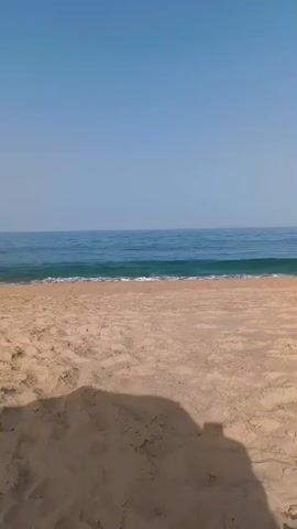 The video opens with a view of a sandy beach and the ocean under a clear blue sky. The camera pans across the sand, revealing footprints and small shadows. In the distance, the waves gently roll onto the shore, and a faint line of land is visible on the horizon. As the video progresses, a bright green umbrella enters the frame, partially obscuring the view of the sky and the sea. A few people can be seen walking along the beach in the distance. The overall atmosphere is peaceful and serene, capturing a typical day at the beach.