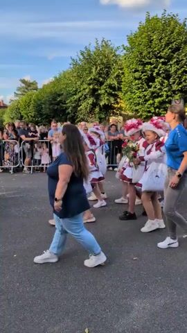 A group of young women in matching red and white costumes with feather hats are marching down a street, holding bouquets of flowers. They are followed by a few adults and a crowd of spectators standing behind a metal barrier. A yellow building with the text "Buttstädter Vollkornbäckerei" is visible in the background. The sky is blue with some clouds, and trees line the street. The overall atmosphere appears to be festive, possibly a parade or a celebration.