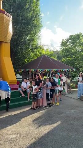 A group of children and an adult woman are standing in a line, as if waiting for something. A man dressed in a costume, holding a microphone, stands to the left of the group. In the background, there is a carousel and trees. The ground is paved.