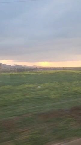 The video shows a landscape viewed from a moving vehicle. A flat field stretches across the foreground, its color a mix of green and dark brown, blurring due to the motion.  In the distance, a range of low mountains can be seen silhouetted against the sky. The sky is predominantly gray with a hint of orange on the horizon, indicating either sunrise or sunset.  The camera is moving, causing the foreground to blur.