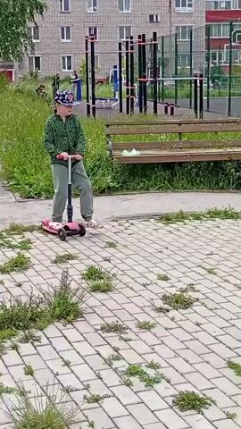 A person wearing a green patterned jacket, grey pants, and a baseball cap rides a pink three-wheeled scooter on a paved walkway. The person is in motion, with their right leg extended backwards. In the background, there is a tall apartment building, a playground with colorful equipment, green grass, trees, and a wooden bench with two people sitting on it. The weather appears to be overcast or cloudy.