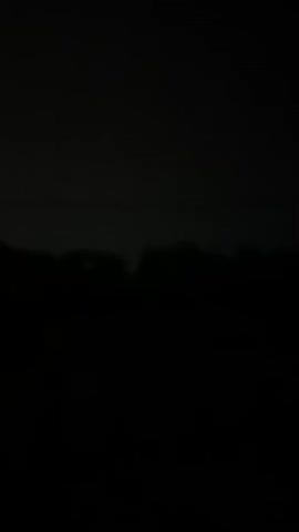 The video displays a dark, blurry scene, likely shot at night or in very low light conditions. The foreground is almost entirely black, obscuring any details. In the middle ground, there is a dark silhouette of what appears to be trees or a treeline. Above the treeline, a faint horizontal line is visible, possibly a power line or a distant structure. The sky is a dark gray, with a subtle gradient suggesting it might be cloudy or overcast. There are no discernible people, objects, or actions taking place. The camera appears to be stationary.