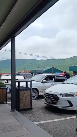 A short video clip shows a view from a covered outdoor seating area of a restaurant or cafe. The seating area has a grey tiled floor and black metal tables and chairs. A retractable awning covers the seating area. Beyond the seating area, there is a parking lot with several cars. In the background, there are mountains and a cloudy sky. Several people are visible in the clip. One person is walking past the seating area, and two people are sitting at a table inside the cafe. Another person is standing near a car in the parking lot. The video appears to be taken during the daytime.