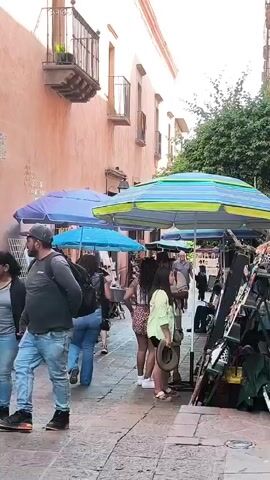 The video shows a bustling street scene in what appears to be a tourist area. People are walking and browsing through merchandise displayed under colorful umbrellas on either side of a cobblestone street. The architecture features tall, peach-colored buildings with wrought-iron balconies. The street is narrow and crowded with vendors and shoppers.