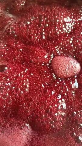 The video shows a close-up, top-down view of a red, bubbly liquid being heated in a pan. Bubbles rise to the surface and pop, creating foam and ripples. The liquid appears to be thick and viscous, possibly a sauce or jam, with a rich, deep red color. The lighting is bright, highlighting the texture and movement of the liquid. There are no people or other objects visible in the frame.