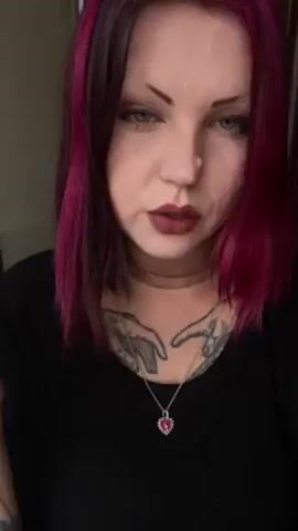 A woman with short, dark purple hair, dark red lipstick, and pale skin inhales and exhales a cigarette. She is wearing a black top and a silver pendant. She has heavy tattoos on her hand and neck. The background appears to be a bathroom.