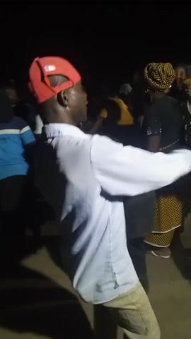 The video shows a group of people dancing at night. Most are only partially visible. One man is prominent; he wears a red baseball cap, a light blue shirt, and khaki pants. He is turned away from the camera, dancing with arm movements. Other people are nearby, including someone with a backpack labeled "STAR BOY". The lighting is poor and grainy, making it difficult to discern details.