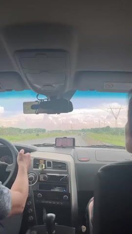 The video shows the interior of a car from the perspective of a passenger. A person's hands are visible on the steering wheel and gear shift. The dashboard and windshield are in focus, with a cloudy sky and distant power lines visible through the windshield. The car appears to be in motion on a paved road surrounded by greenery. A smartphone is mounted on the dashboard.