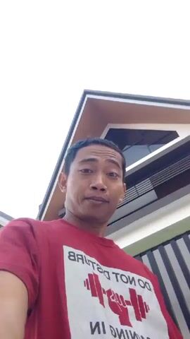 A man with short dark hair and light brown skin is wearing a red t-shirt. He is looking at the camera and smiling. In the background, there is a house with a brown roof and white walls. The sky is overcast. The video is shaky and appears to be a selfie.