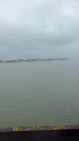 The video, shot from a slightly elevated position, offers a view of a body of water under a cloudy sky. The water is a murky green hue. In the distance, a coastline is visible, dotted with buildings. A black metal structure partially frames the shot on the right. As the video progresses, chains come into view, hanging down in front of the water. The chains appear to be made of heavy metal.