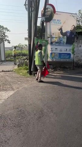 The video shows a street scene with two individuals wearing high-visibility vests. The person in the foreground, facing away from the camera, holds two red flags. They appear to be standing near a pole and a large advertisement billboard in the background. The billboard displays text and images, including a person in a uniform. The environment is outdoors, with a paved road in the foreground and vegetation visible behind the billboard. Sunlight suggests it is daytime. The second individual, also in a high-visibility vest, is visible further down the street, facing away from the camera.