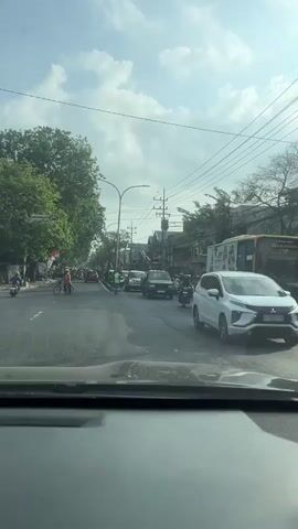 The video shows a street scene captured from inside a vehicle. The road is paved and appears to be in a city, with trees lining the left side and buildings on the right. There are various vehicles on the road, including cars, motorcycles, and a bus. Several people are visible, some on motorcycles and one person standing in the middle of the road holding flags, possibly directing traffic. The sky is cloudy with patches of blue. The overall impression is a busy urban street on a bright, possibly sunny day.