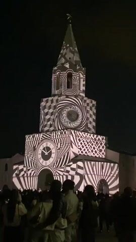 The video shows a tall, multi-tiered, light-colored building at night, with a crowd of people in the foreground watching a light show. The building's exterior is illuminated with changing patterns of light, including abstract geometric designs, words, and shapes. The people in the crowd are mostly silhouetted against the bright building, with some holding up phones to record the event. The background is dark, emphasizing the light display on the building.