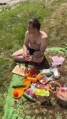 A man is sitting cross-legged on a brown blanket spread on green grass. He is shirtless and wearing dark blue shorts. He holds small red pieces of food, appearing to be tomatoes, in his hands and is eating them. In front of him on the blanket is a picnic spread. There is a wooden cutting board with a knife and sliced tomatoes, orange plates with cucumbers and tomatoes, containers with salad, a package of wet wipes, a white cup, a plate of cooked meat, and a loaf of bread in plastic wrapping. Sunglasses are also visible on the blanket. The background shows more green grass and a sandy path or area. The scene is outdoors on a sunny day.