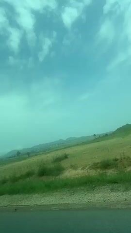 The video shows a view from inside a vehicle looking out at a landscape. The sky is overcast and a greenish-blue color. The landscape consists of green grass along the side of the road, a field of dry grass or dirt beyond that, and rolling hills in the distance. There are a few scattered trees, some with leaves and one that appears bare. The vehicle appears to be a dark-colored SUV. The video is taken from a moving vehicle, indicated by the slight blur and the changing perspective of the scenery. The time stamps suggest the video is very short, only a few seconds long.