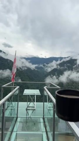 The video begins with a wide shot of a scenic mountainous landscape under a cloudy sky. In the foreground, there is a glass-enclosed observation deck with a small table. A Georgian flag is prominently displayed on the deck. The camera then zooms in on a hand holding a black mug with a golden handle, filled with a light-colored beverage. The hand is holding the mug up as if to take a sip, with the mountainous view in the background. The video concludes by returning to the original wide shot of the observation deck and the landscape.