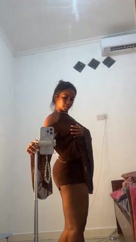 A person is standing in a room in front of a mirror, filming themself with a smartphone attached to a stand. The person is wearing a short, brown dress and a decorative chain on their back. The person initially faces the mirror, and then turns their back to the mirror. They then raise their arms, causing the sleeves of their dress to flare out. The room appears to be a bedroom, with an air conditioning unit on the wall, and a bed and other objects visible in the background. The walls are white.