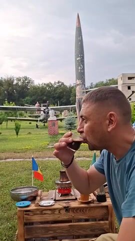 A man in a blue t-shirt is drinking coffee from a small glass cup. He is sitting outdoors at a wooden table with a small stove and flags. In the background, there is a rocket and an airplane, along with some trees and a cloudy sky. The man is holding the cup with his right hand and bringing it to his lips.