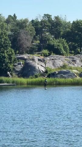 A person in a dark wetsuit is wakeboarding on a lake. The water is blue and calm, with some ripples. In the background, there is a shoreline with tall green grass and trees. Rocky formations are visible behind the trees, and the sky is clear and blue. A wooden dock extends into the water from the left side of the frame, and another person can be seen standing on the dock in the distance.