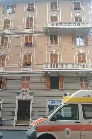 The video shows a street view of a multi-story building with a light brown facade. The building has many windows with wooden shutters, some of which are open. There are also several balconies with white railings. On the ground floor, there are storefronts, including one labeled "BAR GAETTI 4" and another labeled "ORTOPEDIA". An ambulance with red and white markings is parked on the street in front of the building, partially obscuring the storefronts. The ambulance has text on the back that reads "P.A. NERVIESE". The sky is overcast. The video pans slightly to the left, showing more of the building's facade and the street.