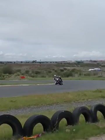 The video depicts a person riding a motorcycle on a racetrack. The rider is wearing a white helmet and riding gear. The racetrack is surrounded by grass, trees, and a few buildings in the distance. There are a few people standing around the track, including a person in an orange vest who appears to be filming. There is also a Coca-Cola truck visible in the background. The weather is cloudy. There are black tires lined along the bottom edge of the screen.